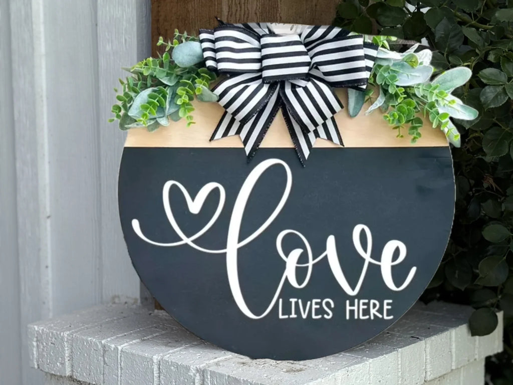A round black sign with white script reads love lives here, decorated with green leaves and a black-and-white striped bow at the top, leaning against a white brick surface.