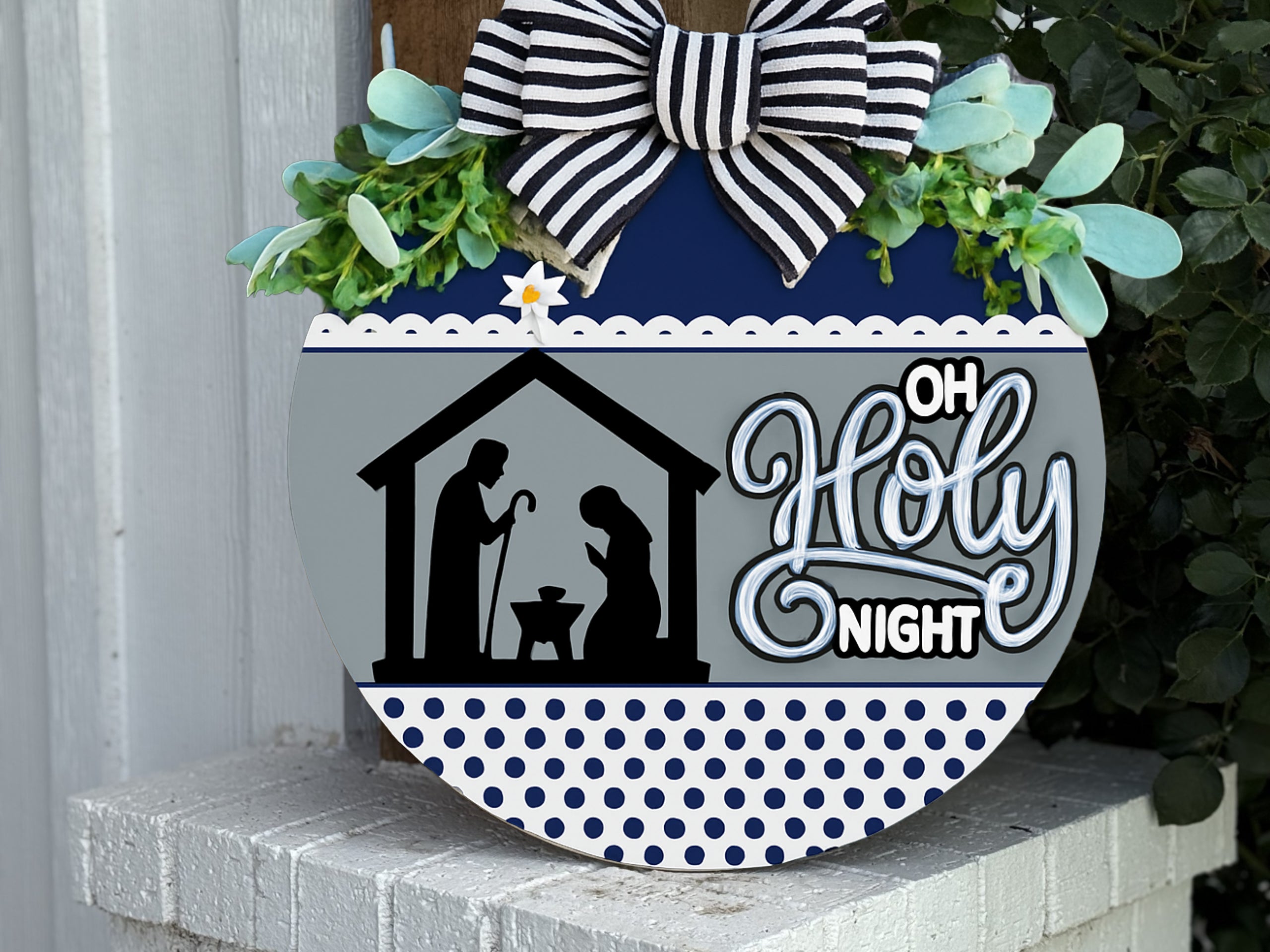 A round sign decorated with greenery and a striped bow features a nativity silhouette, the words Oh Holy Night, and a blue and white polka dot pattern along the bottom.