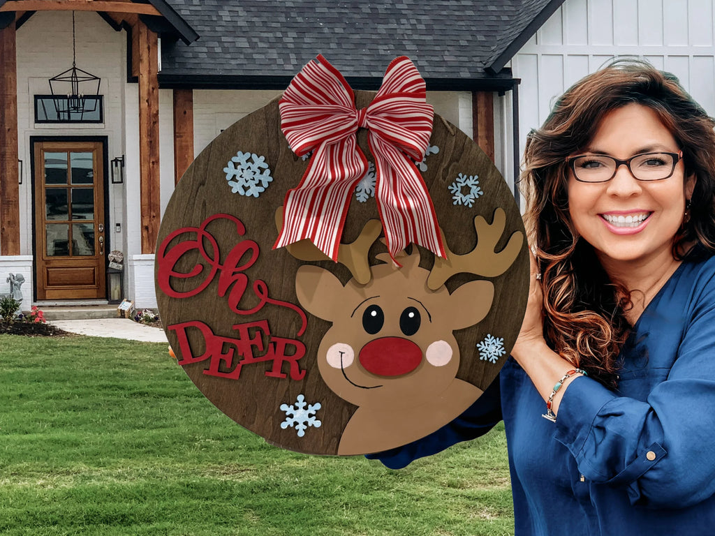 A smiling woman holds the Oh Deer, Rudolph door sign featuring a cartoon reindeer, red and white striped bow, and snowflakes in front of a modern house with white siding and wooden accents.