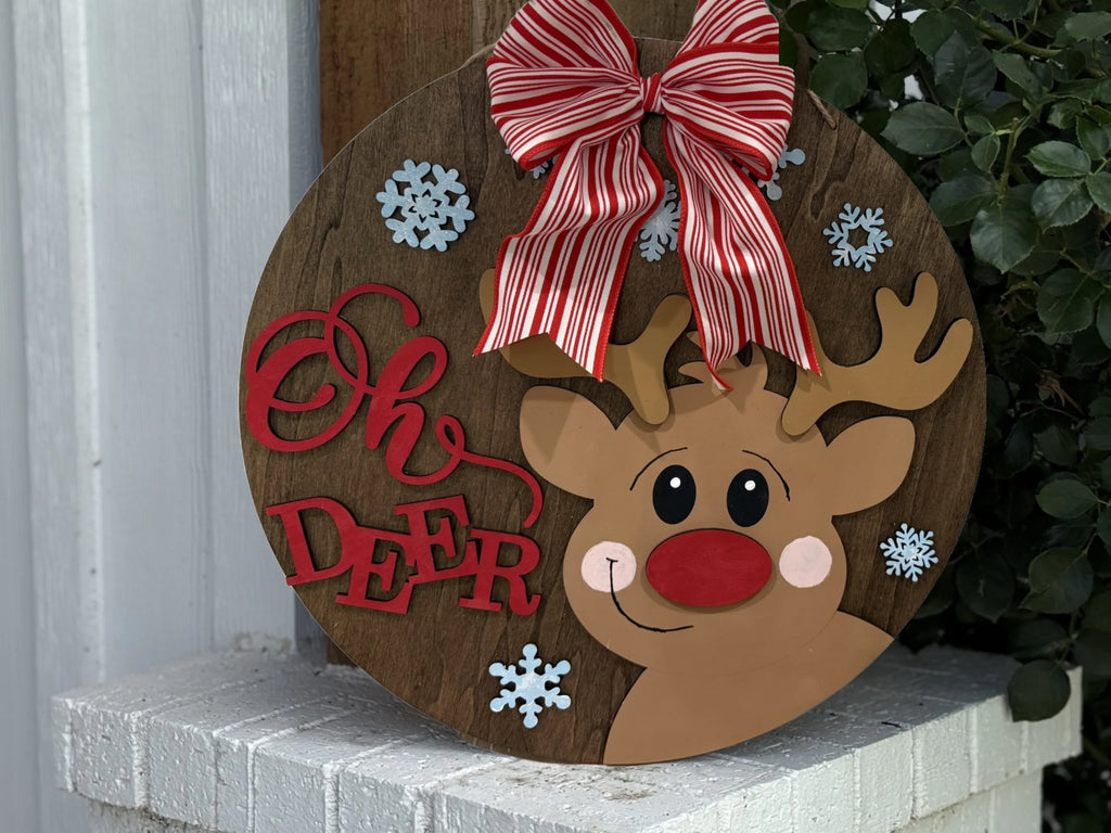 The Oh Deer, Rudolph door sign is a round wooden hanger with a cartoon Rudolph, red-and-white striped bow, snowflakes, and bold red letters. Shown on a white brick surface beside green foliage—perfectly festive decor!.