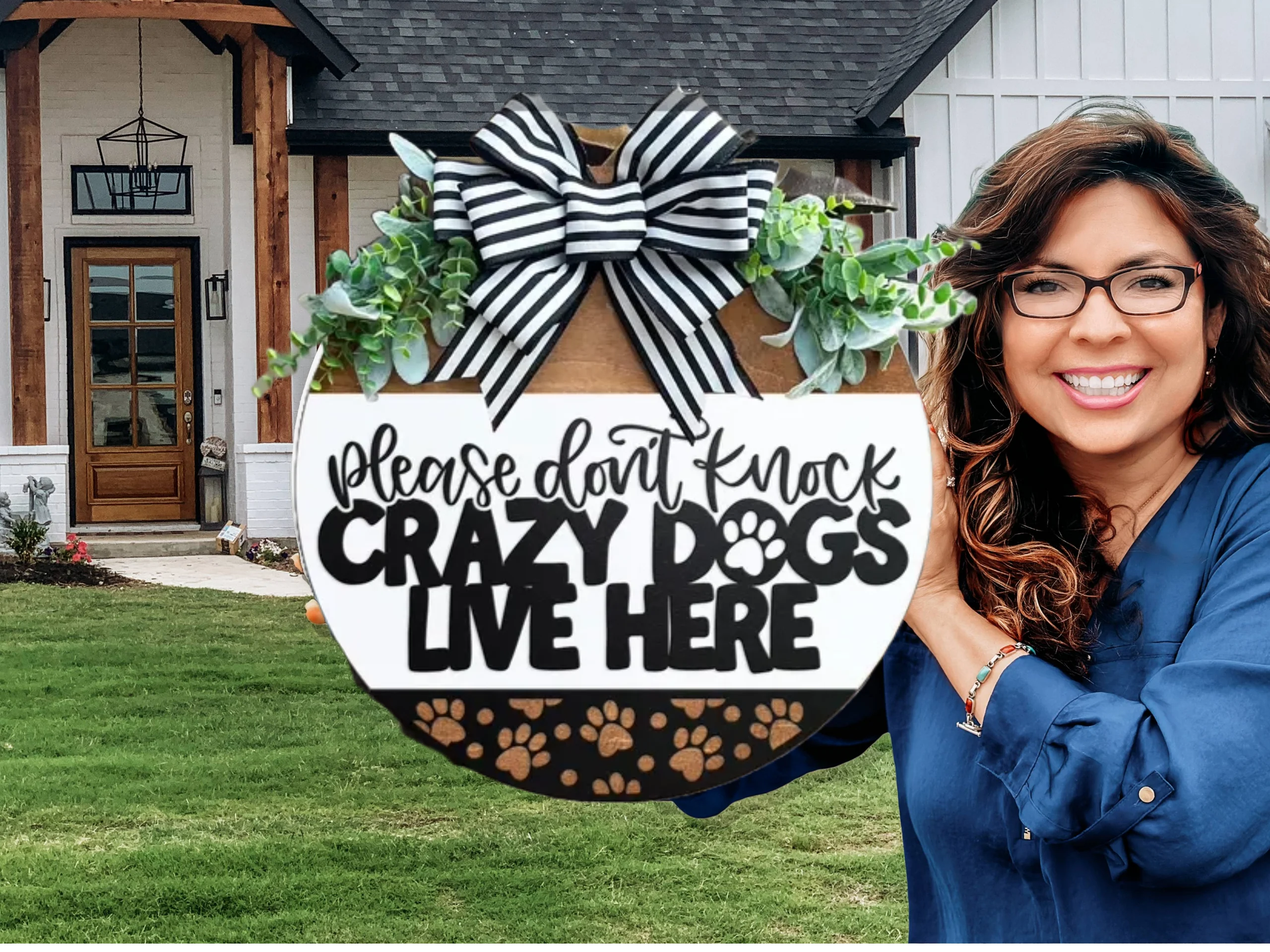 A smiling woman holds a round sign decorated with greenery and a black-and-white striped bow that reads, Please dont knock. Crazy dogs live here. She stands in front of a modern house with a large yard.