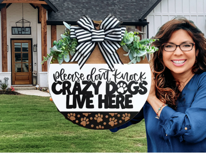 A smiling woman holds a round sign decorated with greenery and a black-and-white striped bow that reads, Please dont knock. Crazy dogs live here. She stands in front of a modern house with a large yard.