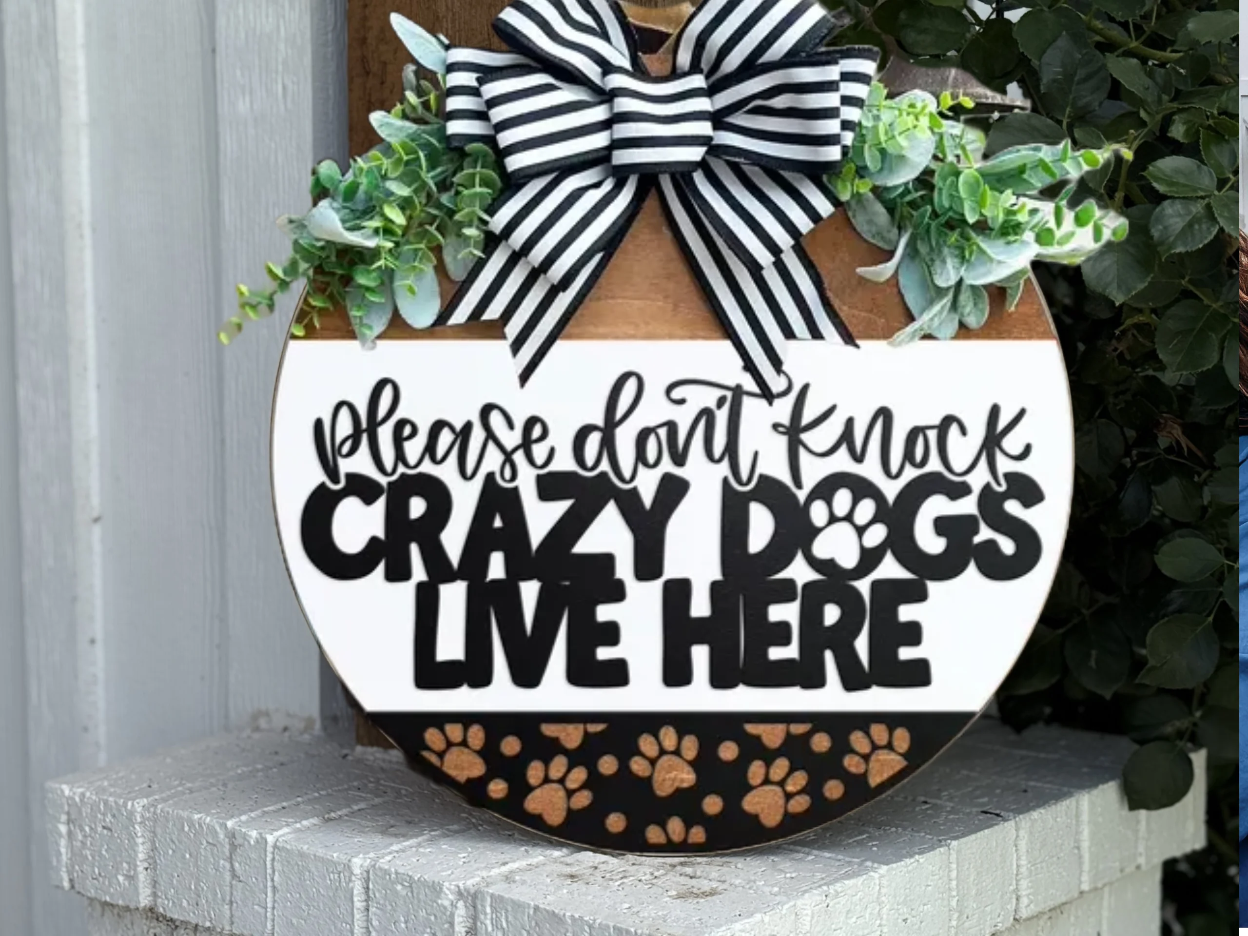 A round door sign with a black and white striped bow, greenery, and the text Please dont knock, crazy dogs live here in bold black letters, with paw prints along the bottom.