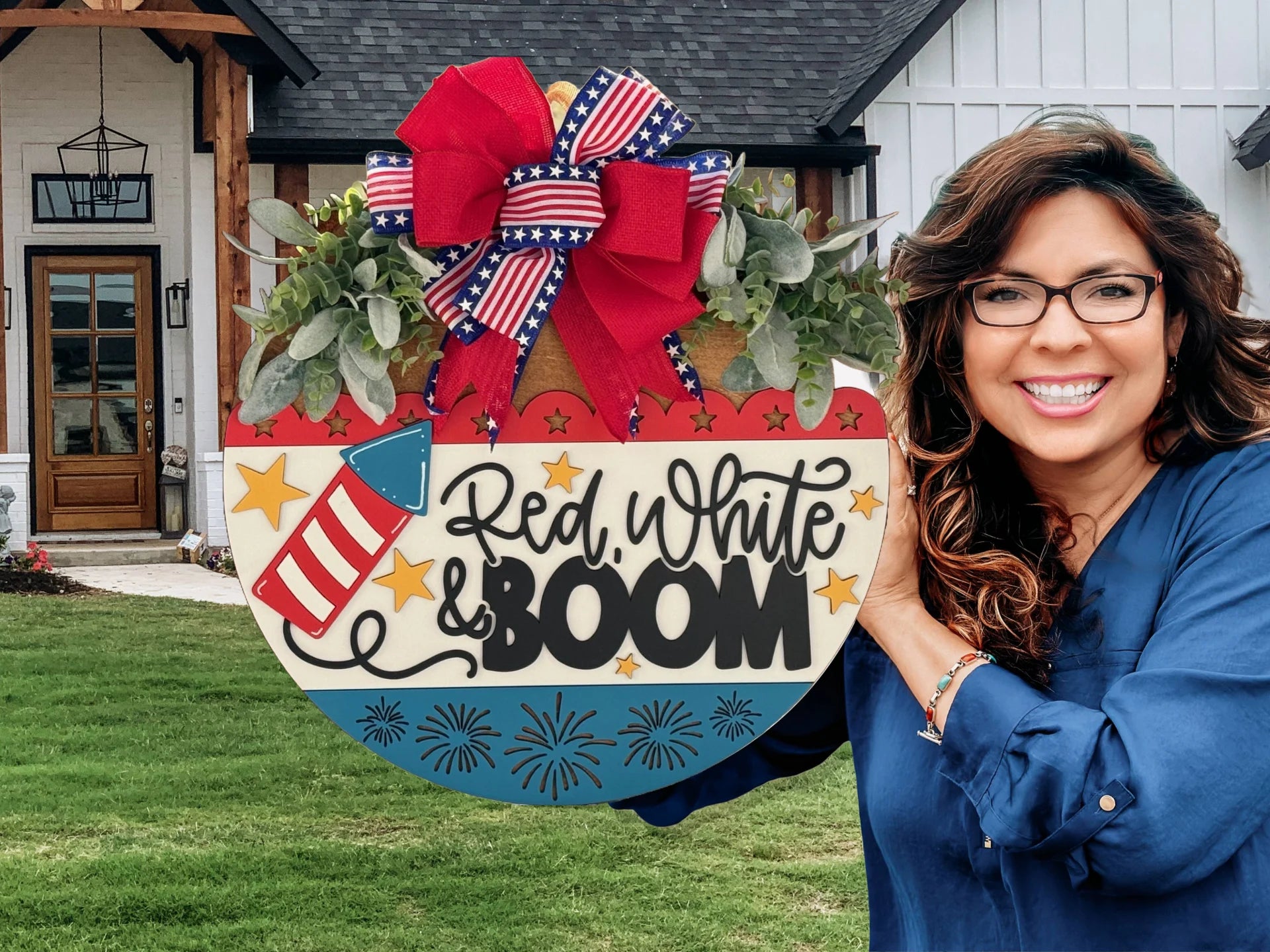 Smiling woman holding a patriotic door hanger featuring a red, white, and blue color scheme with stars, fireworks, and a rocket. The text Red, White & BOOM is displayed prominently. She stands in front of a modern farmhouse with a green lawn and wooden front door.