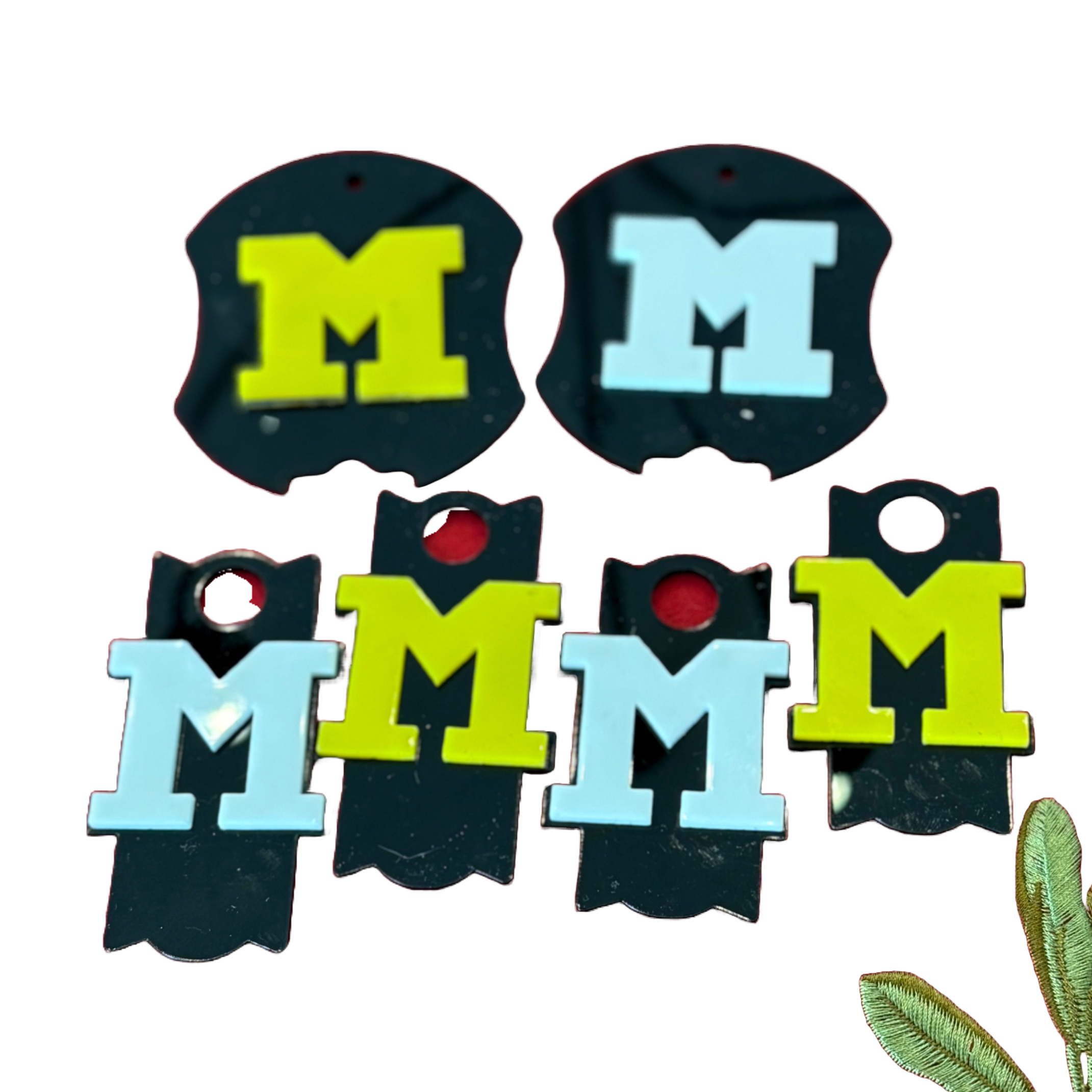 A set of five black cup toppers, each with a large letter M in yellow, light blue, or vibrant green. Arranged in two rows, the top row has two shield-shaped keychains, and the bottom row has three rectangular ones. A small partial view of a leaf is in the bottom right corner.