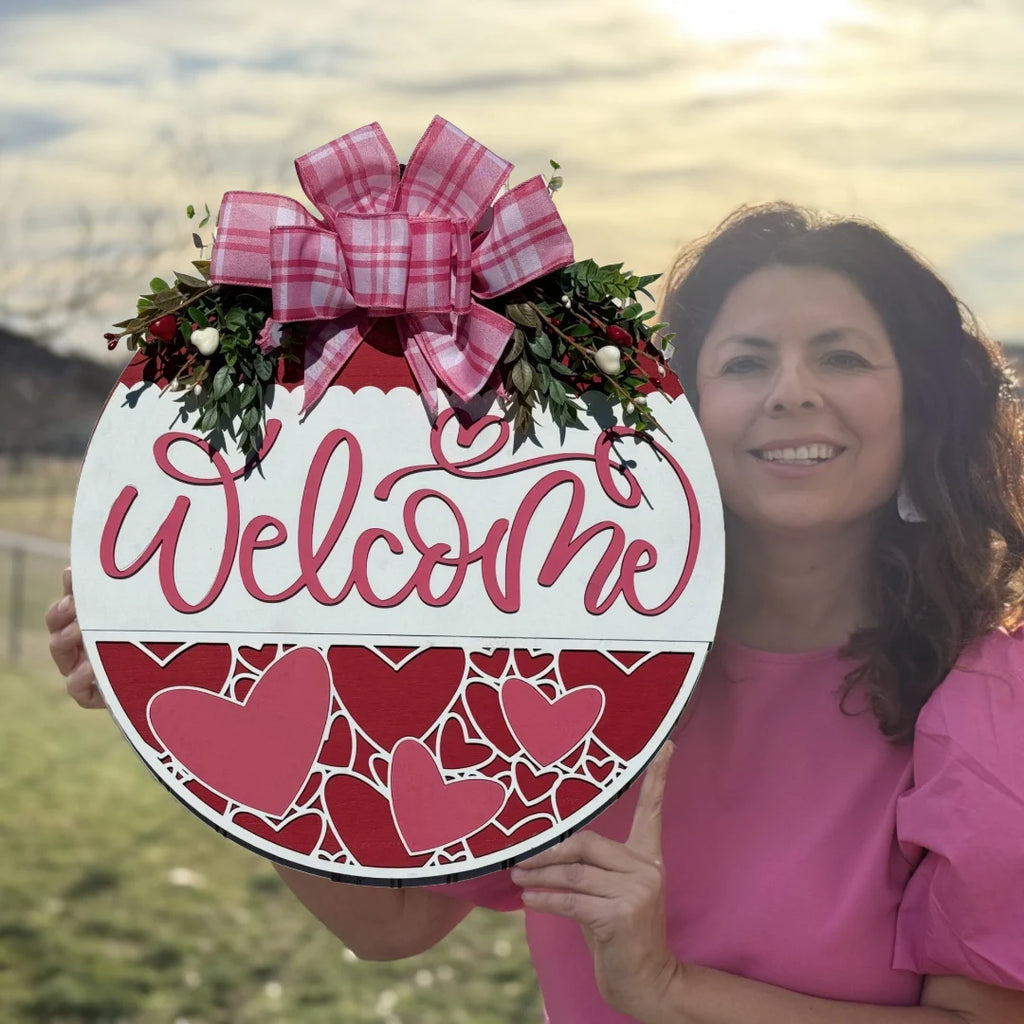 In a grassy field under a cloudy sky, a woman in a pink blouse holds an enchanting Valentine Door Sign. The large circular piece says Welcome in pink cursive on white, decorated with red hearts and a pink plaid bow—its the ideal Wood Door Sign for Valentines Day.