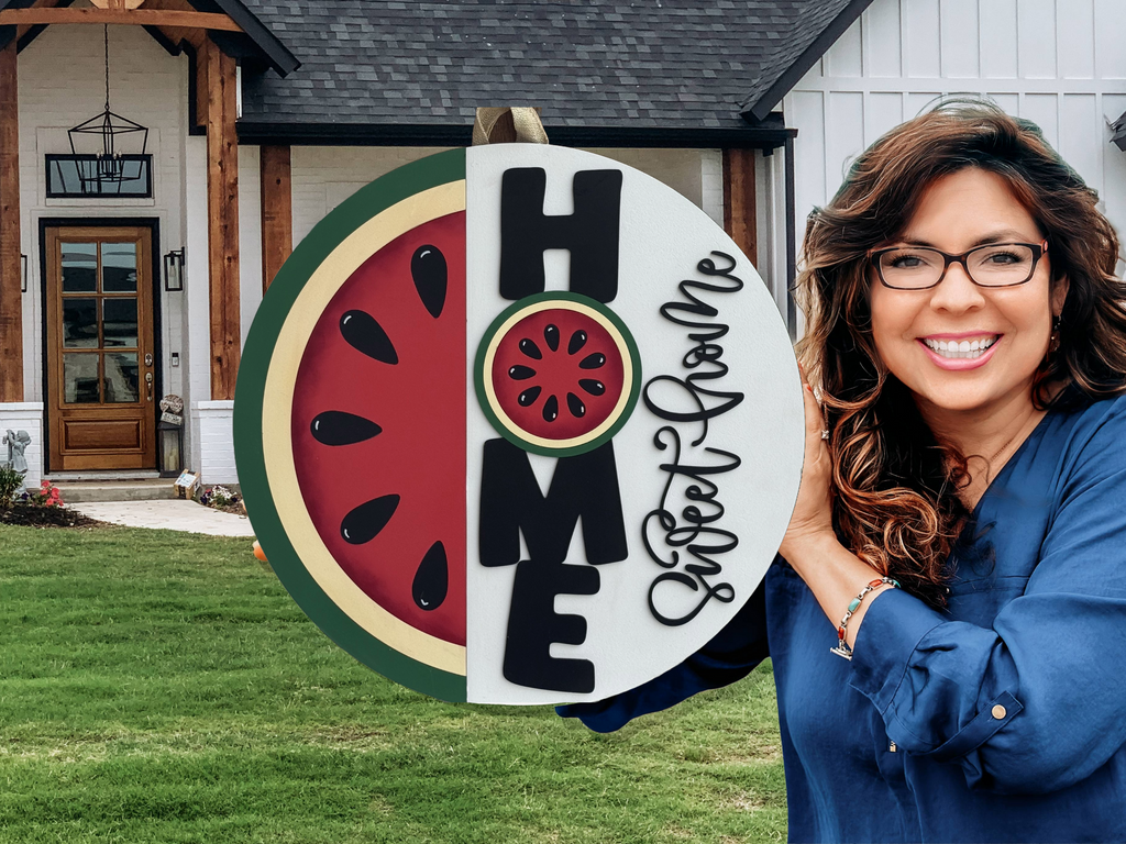 A smiling woman with long brown hair and glasses holds a round Home Sweet Home sign with a watermelon design in front of a modern farmhouse.