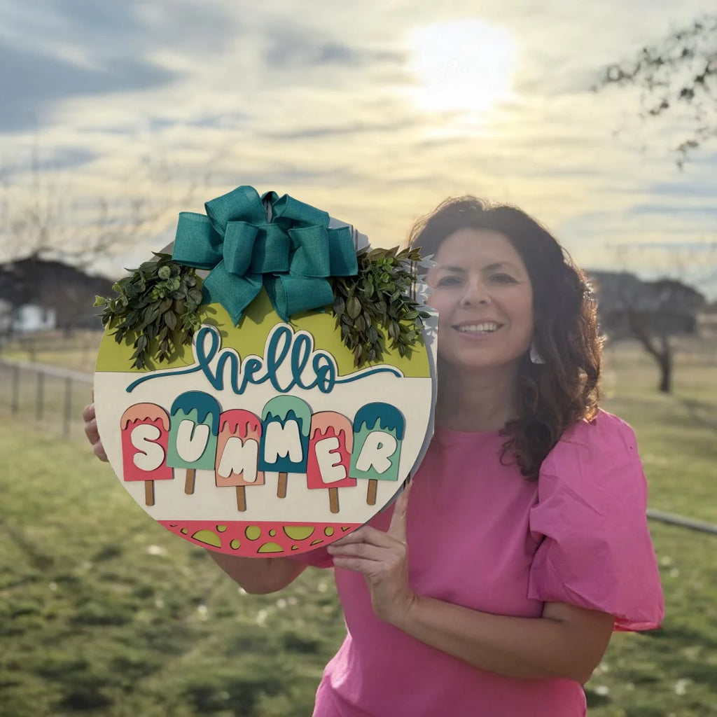 A woman in a pink blouse smiles while holding the Home Sweet Home Door Sign, a decorative piece resembling a door wreath that reads hello SUMMER with colorful popsicles, adorned with a green bow and foliage. Sunlight filters through the cloudy sky over grassy fields with houses in the background.