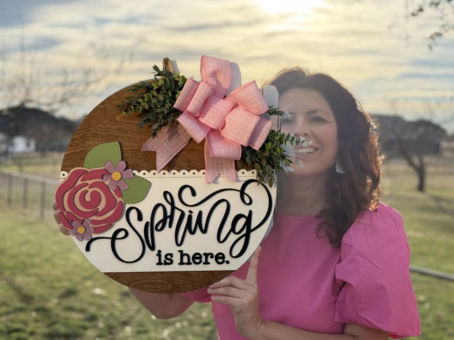 A woman in a pink blouse shows off the Spring Is Here Door Sign, Spring Door Wreath, featuring flowers, greenery, and a pink bow. She stands among bare trees and green grass in a sunny outdoor setting.