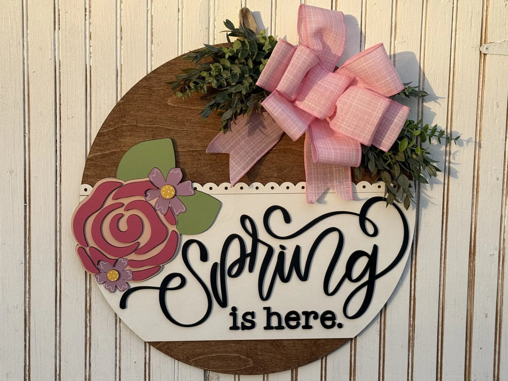 The Spring Is Here Door Sign features a circular wooden design with spring is here in black cursive on a striped background. Adorned with green leaves, a pink bow, vibrant flowers on the left, and a scalloped white edge separating sections, it resembles a delightful spring wreath.