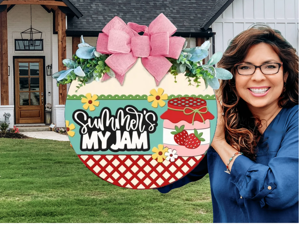 A smiling woman with glasses holds a colorful round sign decorated with a pink bow, flowers, and strawberries. The sign reads Summers My Jam. She stands outside in front of a modern farmhouse-style home.