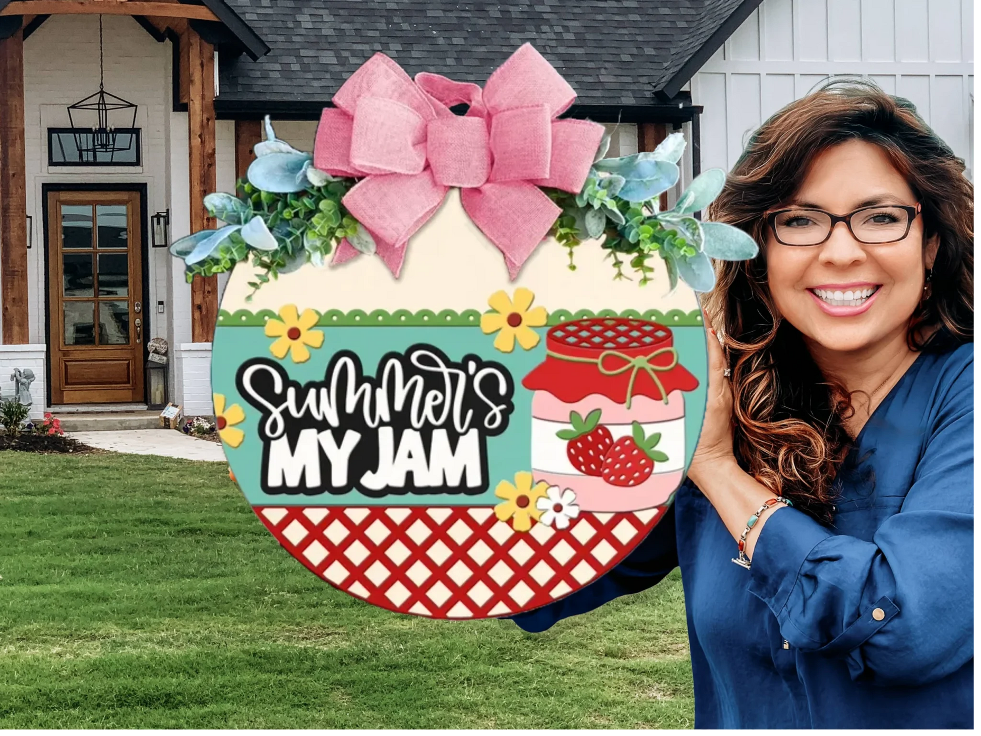 A smiling woman with glasses holds a colorful round sign decorated with a pink bow, flowers, and strawberries. The sign reads Summers My Jam. She stands outside in front of a modern farmhouse-style home.