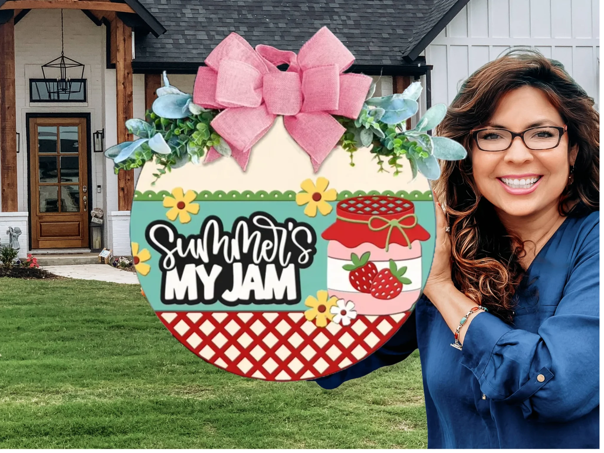 A smiling woman with glasses holds a colorful round sign decorated with a pink bow, flowers, and strawberries. The sign reads Summers My Jam. She stands outside in front of a modern farmhouse-style home.
