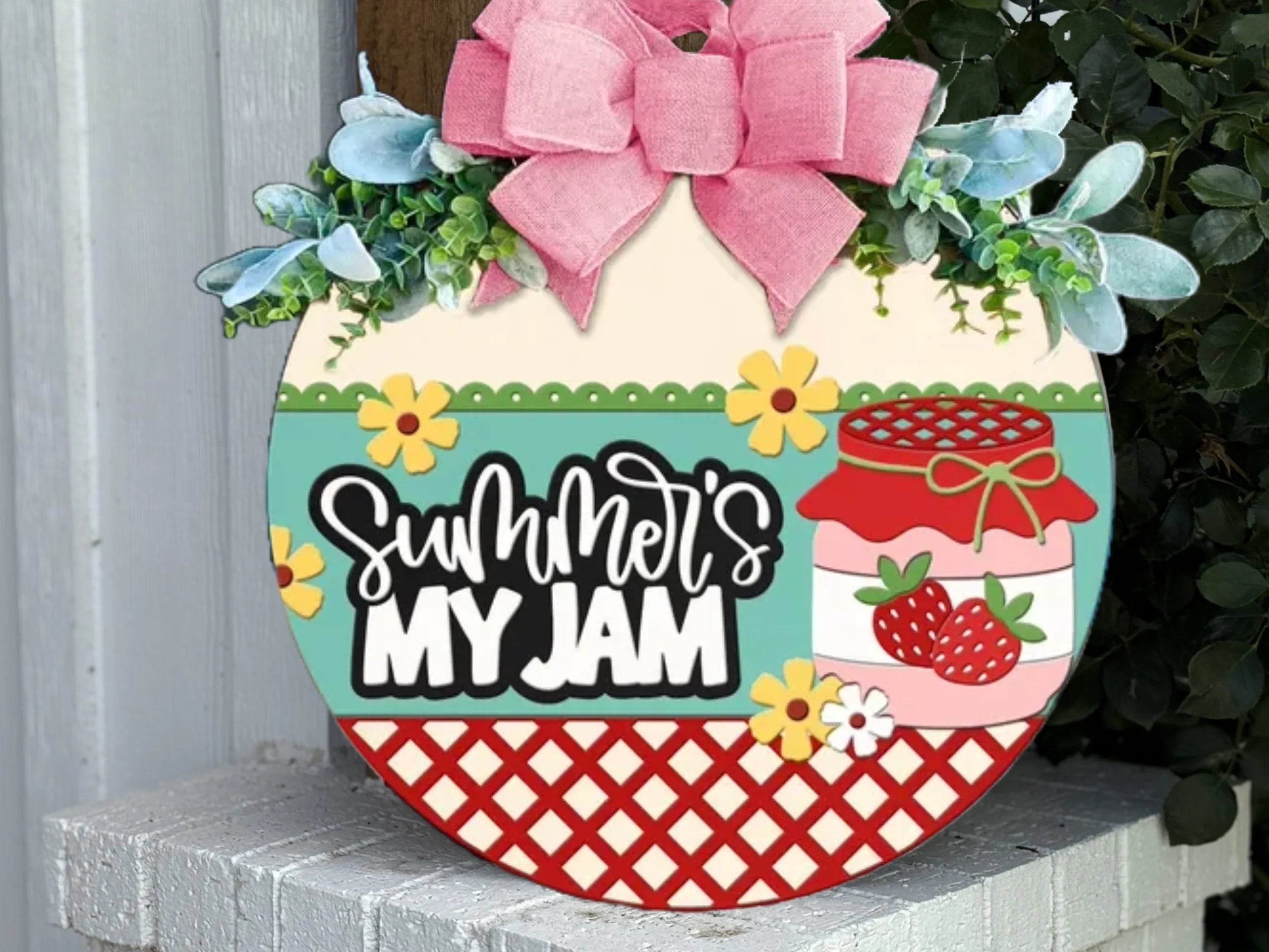 A round decorative sign with a pink bow, greenery, and flowers reads “Summer’s My Jam” next to a jar of strawberry jam. Red lattice and colorful accents decorate the bottom half of the sign.