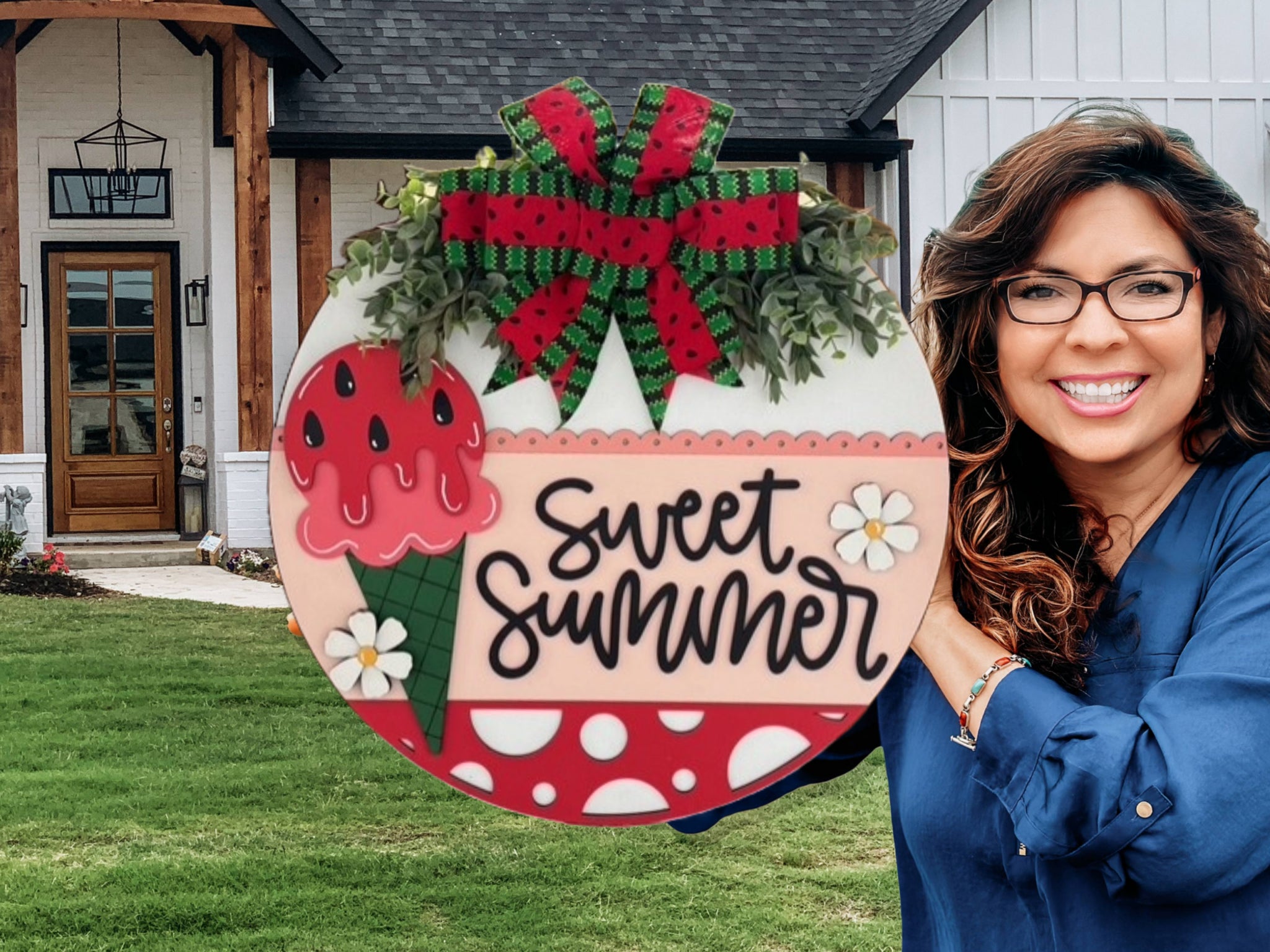 A smiling woman with wavy hair and glasses holds a round, colorful sign that says Sweet Summer with watermelon and ice cream decorations, standing in front of a house with a green lawn.