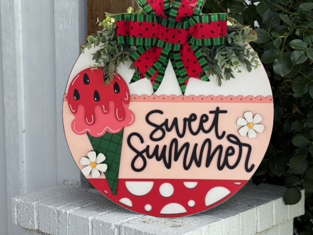A round sign decorated with a green and red bow, faux greenery, and a painted ice cream cone with watermelon-style melting ice cream. The words Sweet Summer are written in black script. White flowers and polka dots decorate the sign.