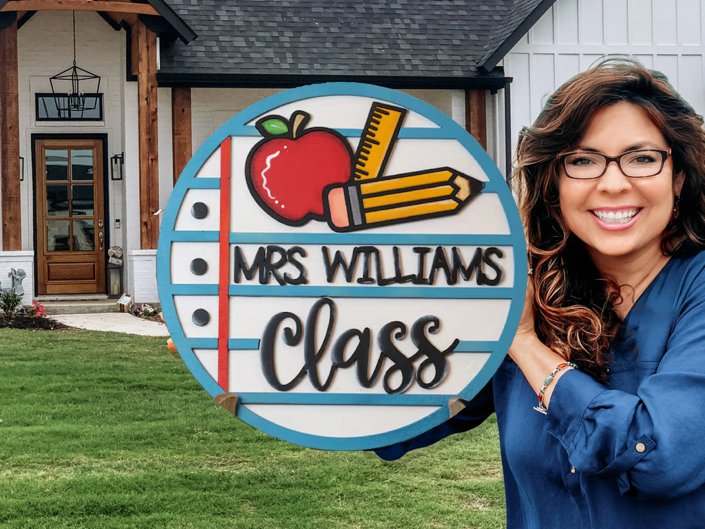 A smiling woman with long brown hair and glasses holds a sign featuring an apple, two pencils, and the words Mrs. Williams Class in front of a house with a green lawn.