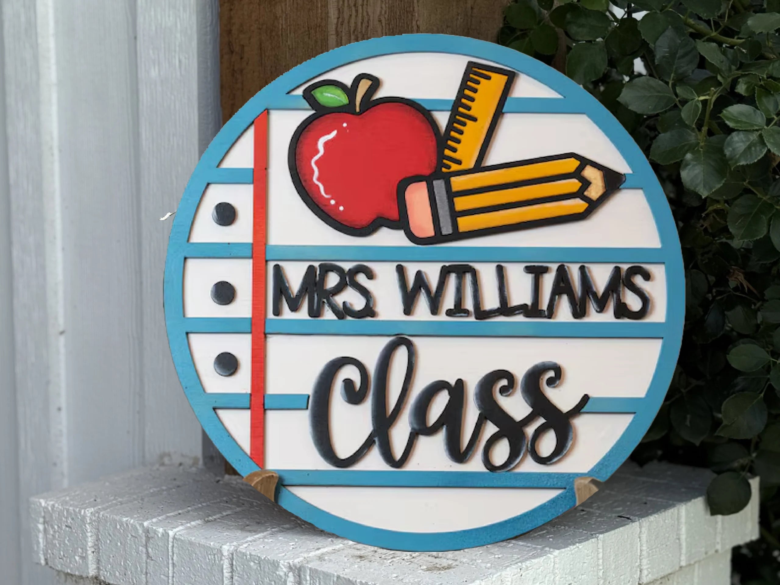 A round sign designed like notebook paper features an apple, pencil, and ruler, with text reading Mrs. Williams Class in a playful font, displayed outdoors near greenery and a white brick surface.