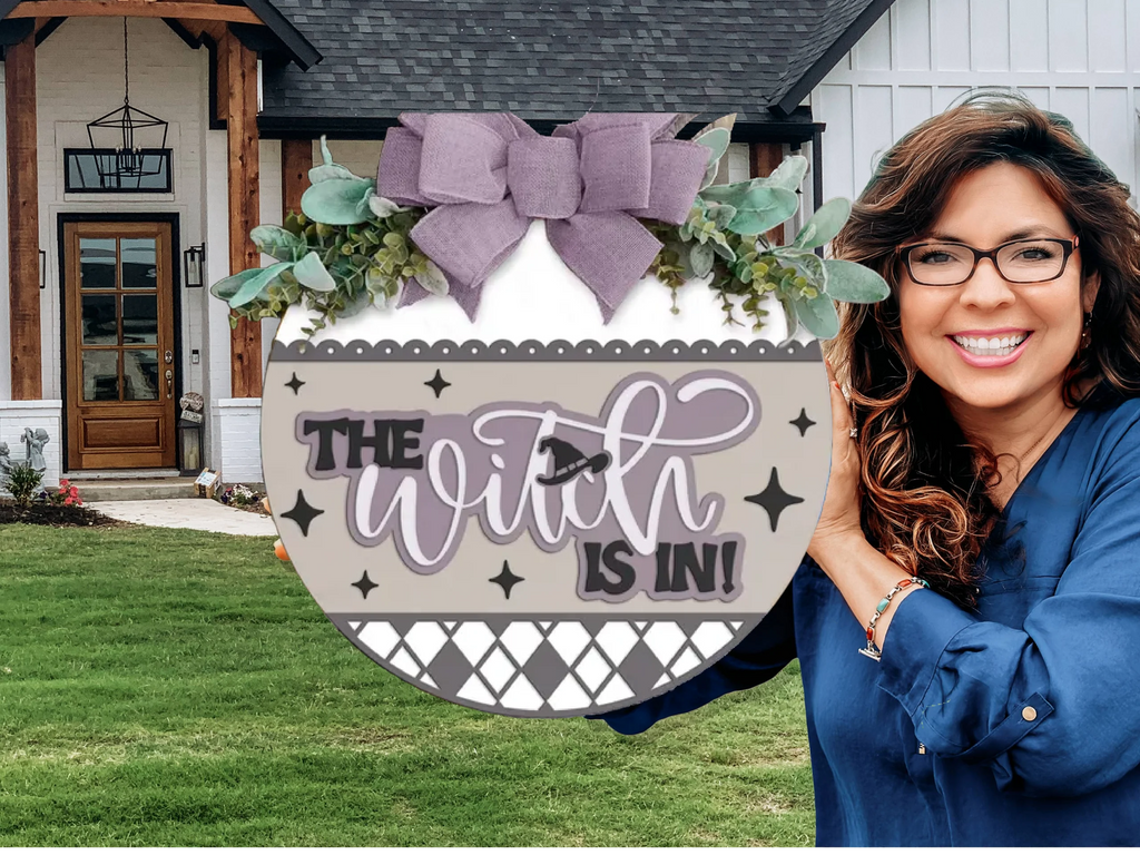 A smiling woman with long brown hair and glasses holds a round decorative sign reading The Witch Is In! in front of a white house with a wooden door and green lawn. The sign is adorned with a large purple bow and greenery.