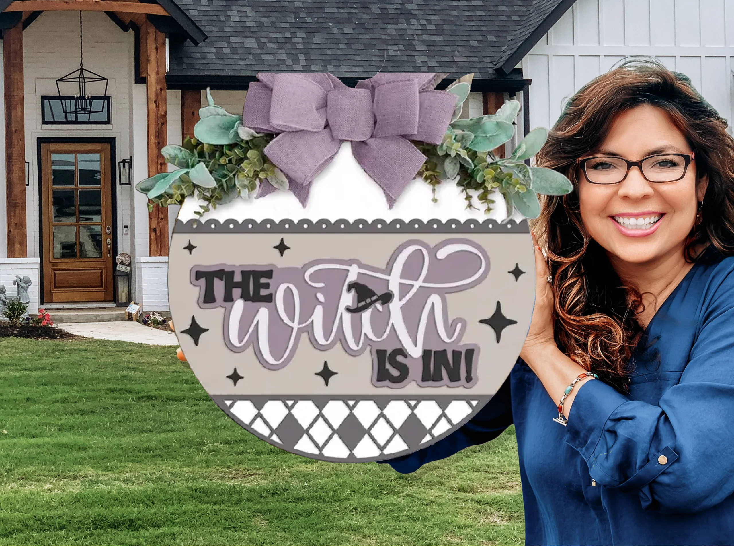 A smiling woman with long brown hair and glasses holds a round decorative sign reading The Witch Is In! in front of a white house with a wooden door and green lawn. The sign is adorned with a large purple bow and greenery.