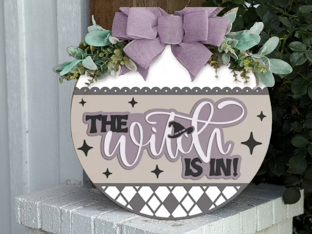 A decorative round sign with greenery and a lavender bow reads The Witch Is In! in playful script, featuring stars and diamond patterns, displayed on a white porch with plants nearby.