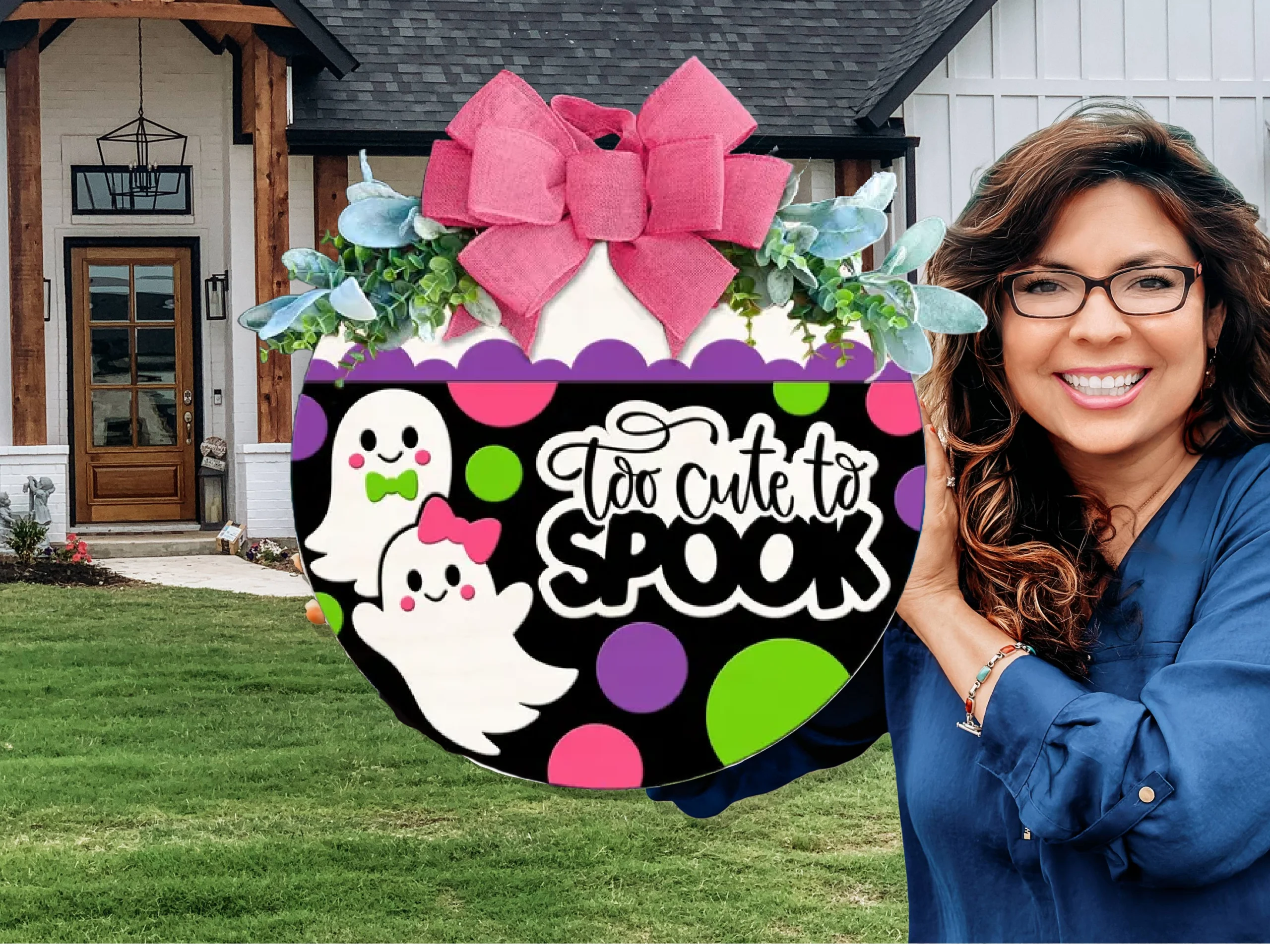 A smiling woman in glasses holds a round Halloween sign with two cartoon ghosts, colorful polka dots, a pink bow, and the text “Too cute to spook” in front of a house with a green lawn.