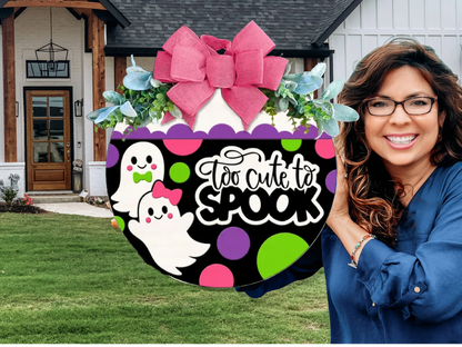 A smiling woman in glasses holds a round Halloween sign with two cartoon ghosts, colorful polka dots, a pink bow, and the text “Too cute to spook” in front of a house with a green lawn.