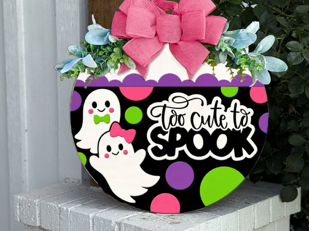 A decorative Halloween sign with two smiling cartoon ghosts, colorful polka dots, the phrase Too cute to spook, a large pink bow, and green leaves, displayed on a white brick shelf.