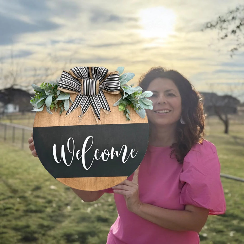 A woman in a pink blouse holds the Welcome Door Sign, featuring a striped bow and greenery. It showcases a black stripe with Welcome in white cursive. She stands on grass, framed by a cloudy sky and bare trees.
