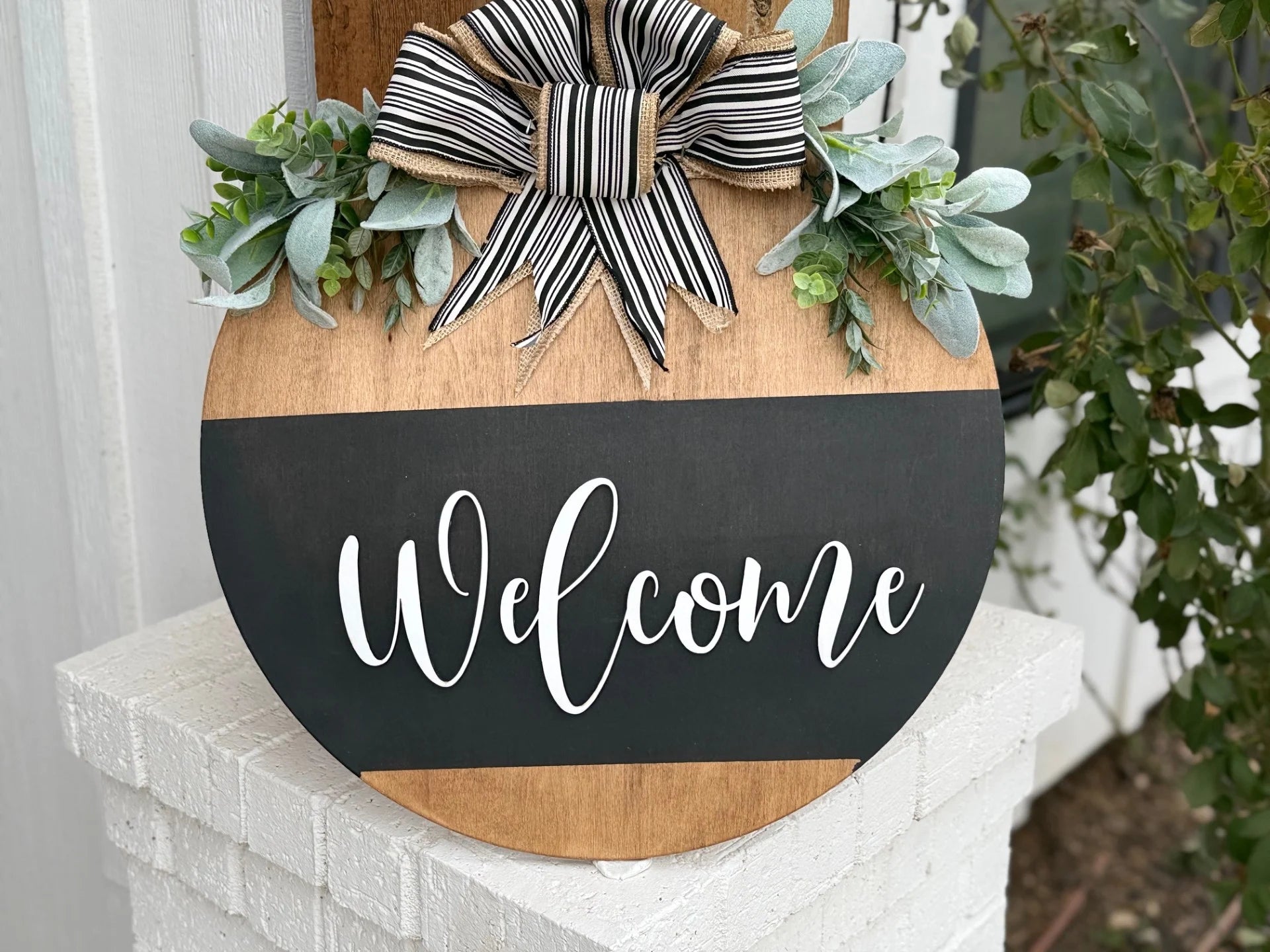 A Welcome Door Sign features a round wooden design with a black chalkboard-style center and Welcome in white script, adorned with a black and white striped bow and surrounded by lush artificial greenery, elegantly displayed on a white brick ledge near a window.