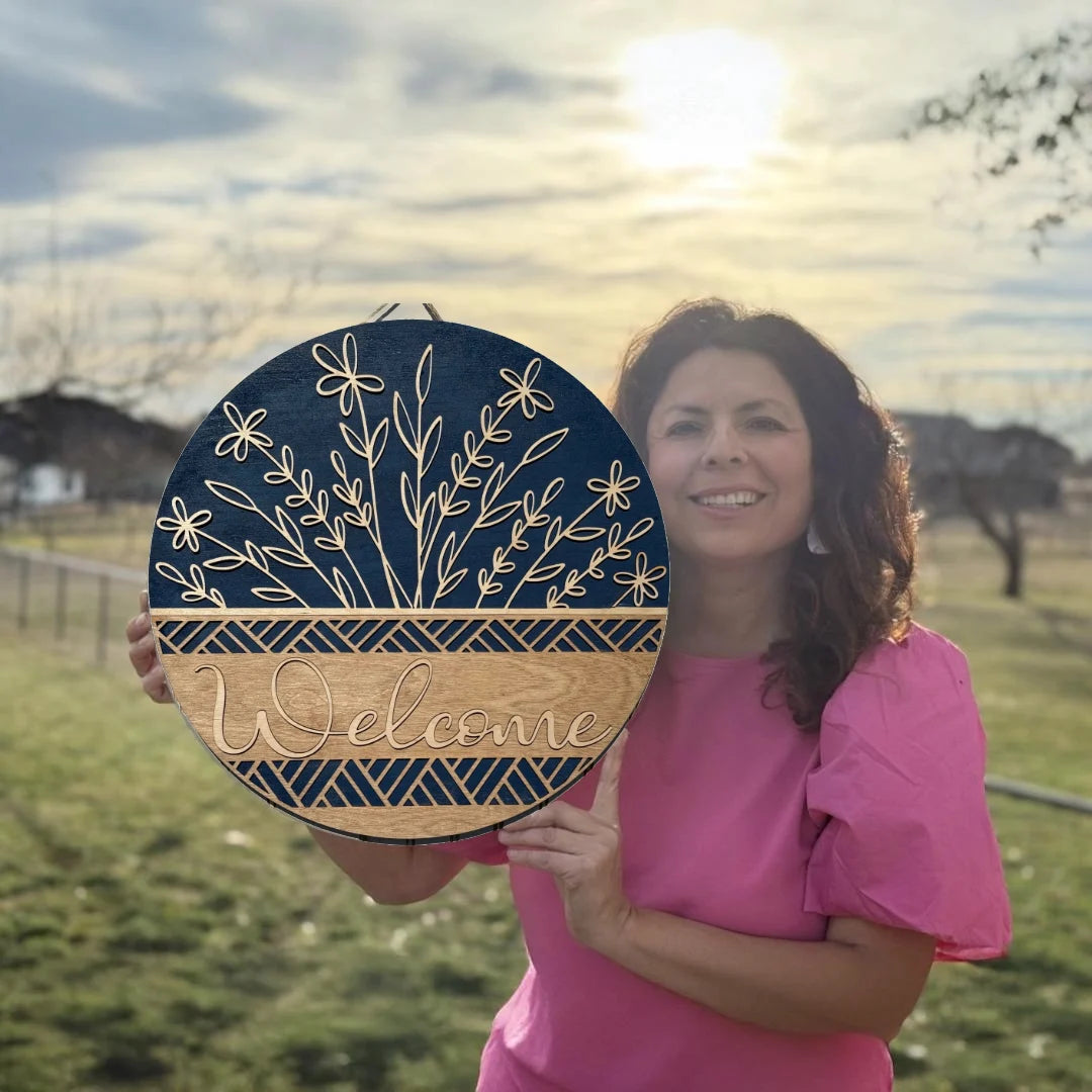 A woman in a pink shirt holds a round wooden sign with floral patterns and the word Welcome engraved on it. She stands outdoors against a cloudy sky with the sun shining brightly behind her, surrounded by a grassy field and fences in the background.