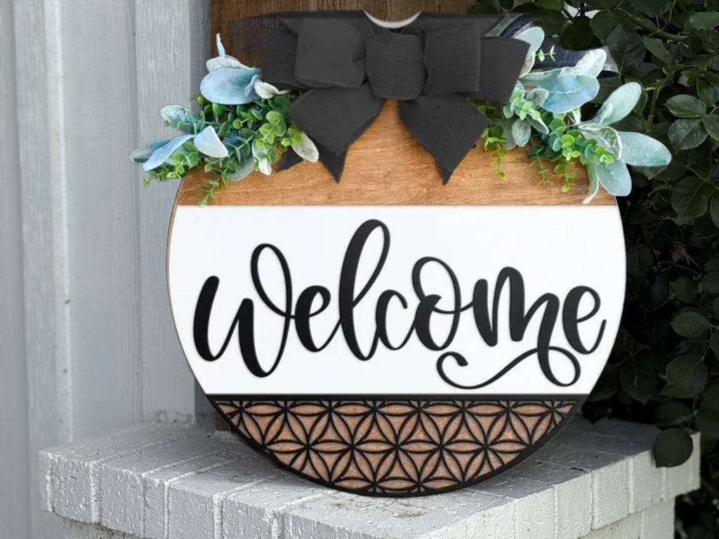 A round wooden welcome sign decorated with green foliage, white flowers, a large black bow on top, and a geometric pattern at the bottom, hanging on a white brick wall.