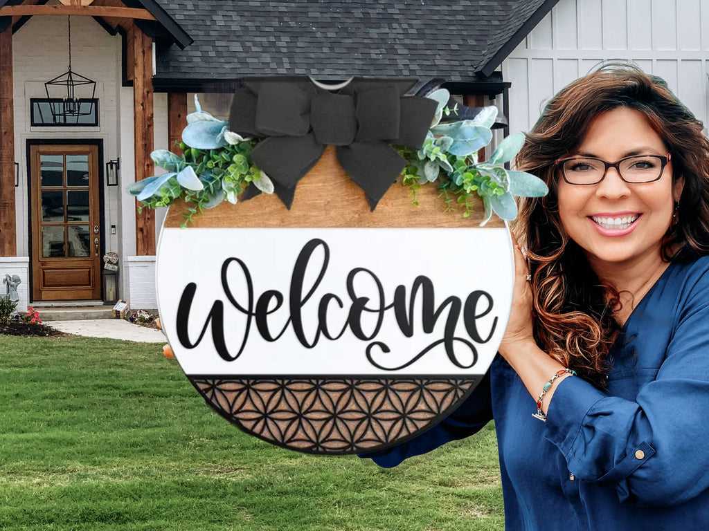 A smiling woman with wavy hair and glasses stands outside a house, holding a round wooden sign decorated with greenery, a black bow, and the word Welcome written in script.