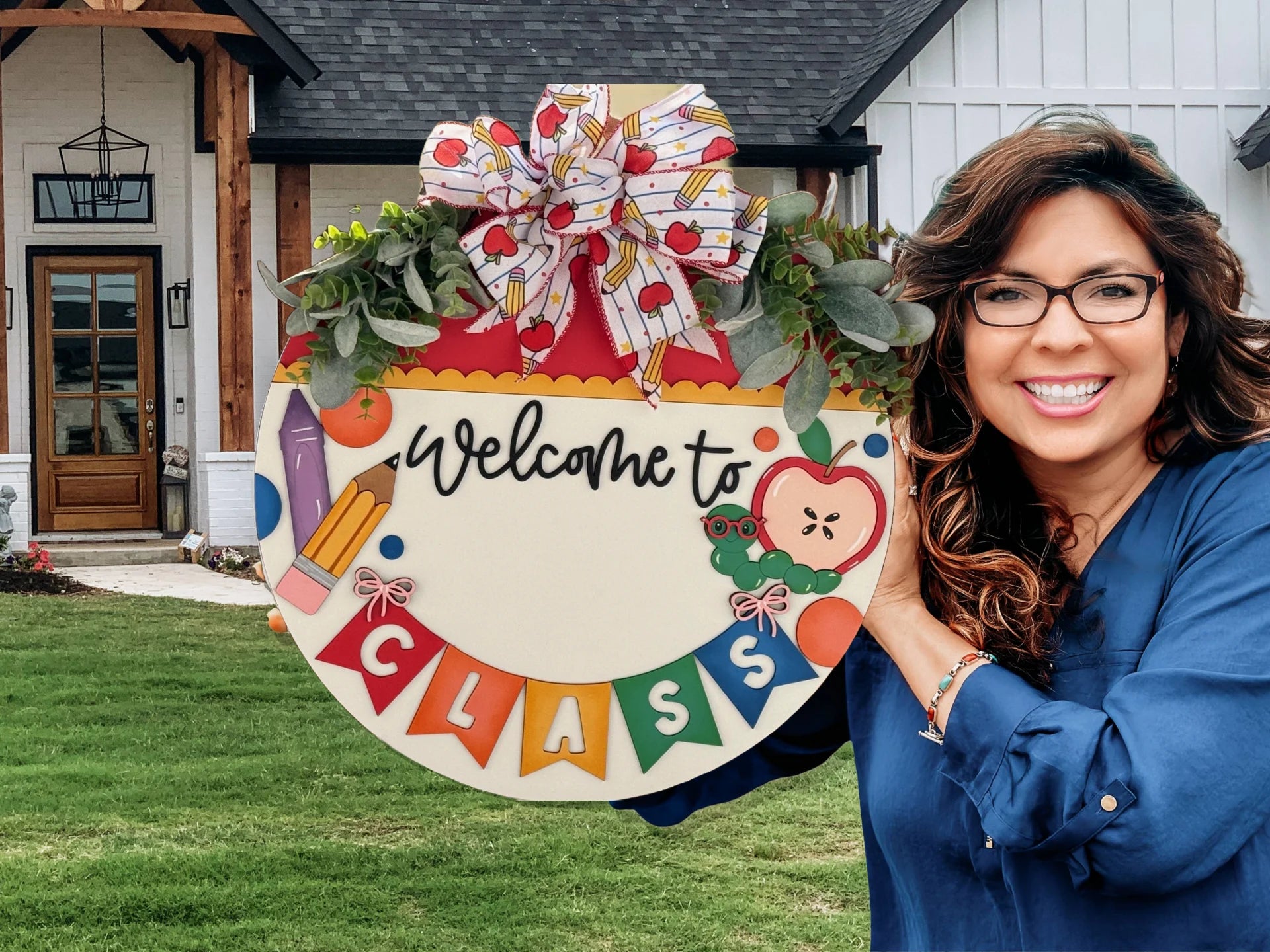 A woman stands smiling outside near a house, holding a round decorative sign that reads Welcome to Class. The sign is adorned with colorful school-themed illustrations, a bow, and greenery. She is wearing glasses and a blue blouse.