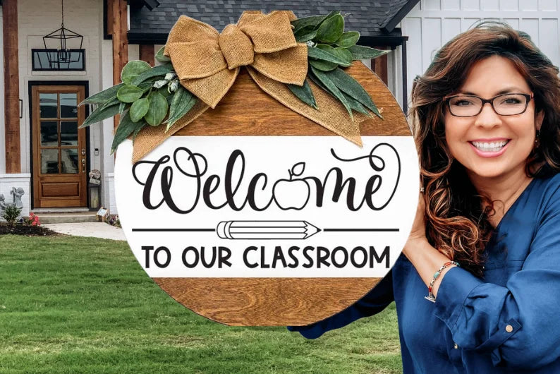 A smiling woman holds a round wooden sign decorated with greenery and a burlap bow. The sign reads “Welcome to our classroom” with an apple and pencil illustration. A house with a porch is visible in the background.