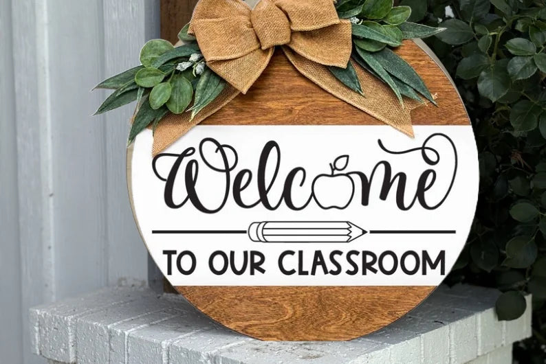 A round wooden sign with a bow and greenery reads Welcome to our Classroom in decorative lettering, with an apple and pencil illustration, displayed on a white brick ledge.