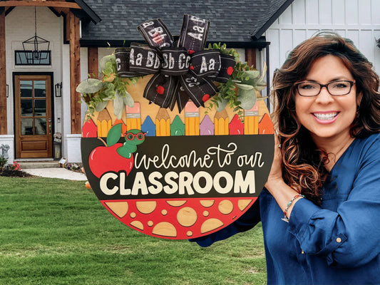 Woman in a blue shirt smiling and holding a colorful Welcome to Our Classroom sign. The sign has a pencil, apple, and book design with a black ribbon featuring the alphabet. She stands outside a house with wooden accents, a glass door, and manicured lawn.