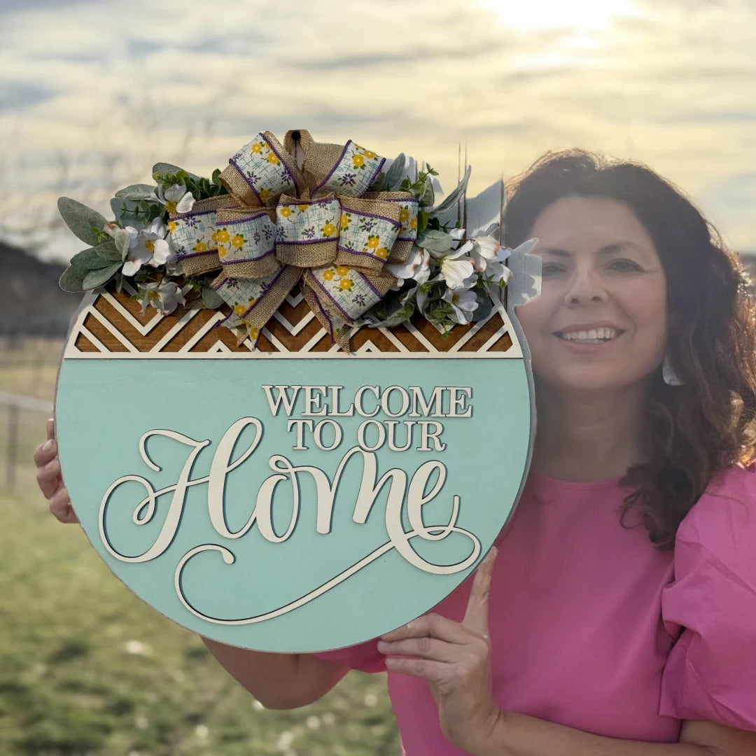 A woman in a pink blouse holds a circular door wreath featuring greenery, flowers, and a decorative bow. The sign reads “Welcome To The Fuster Cluck” in elegant script. A cloudy sky with the sun visible and a blurred outdoor landscape sets the scene.