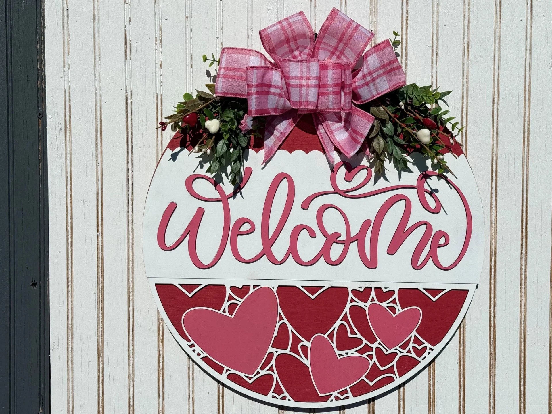 The Welcome Valentines Door Sign, a round, decorative piece on a white striped wall, features Welcome in pink script. Its adorned with a pink plaid bow and greenery atop, complemented by red and pink heart designs below.