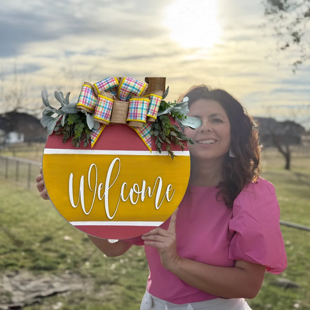 A woman in a pink blouse holds a vibrant Welcome Door Sign, Welcome Wreath, Welcome Door Hanger, adorned with a bow and greenery. The setting sun warms the partly cloudy sky as she stands surrounded by trees in an outdoor grassy area with a fenced yard subtly framing the scene.