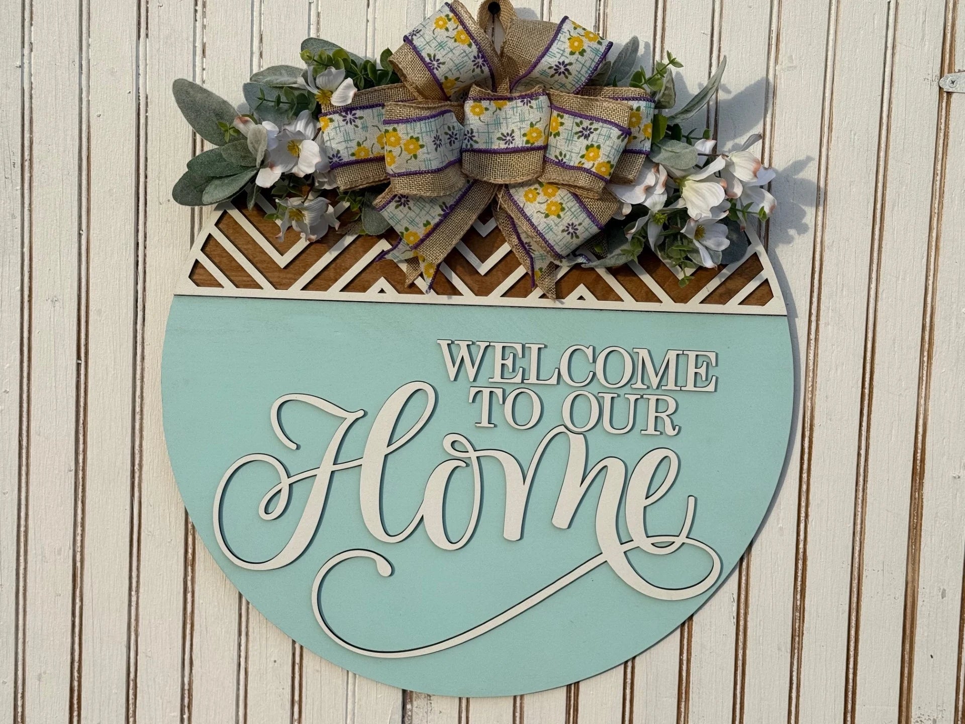 A round sign, titled Welcome To The Fuster Cluck, showcases elegant white script on a light blue background. The top part features a geometric wood design with a large bow decorated with yellow flowers, green and white leaves, all set against a wood-textured backdrop.