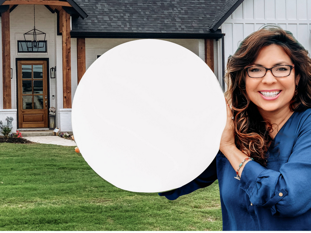A smiling woman with wavy hair and glasses, in a blue shirt, holds a large Laser Cut Wood Round (1/4 inch thick) in front of a modern house with a porch and dark roof.