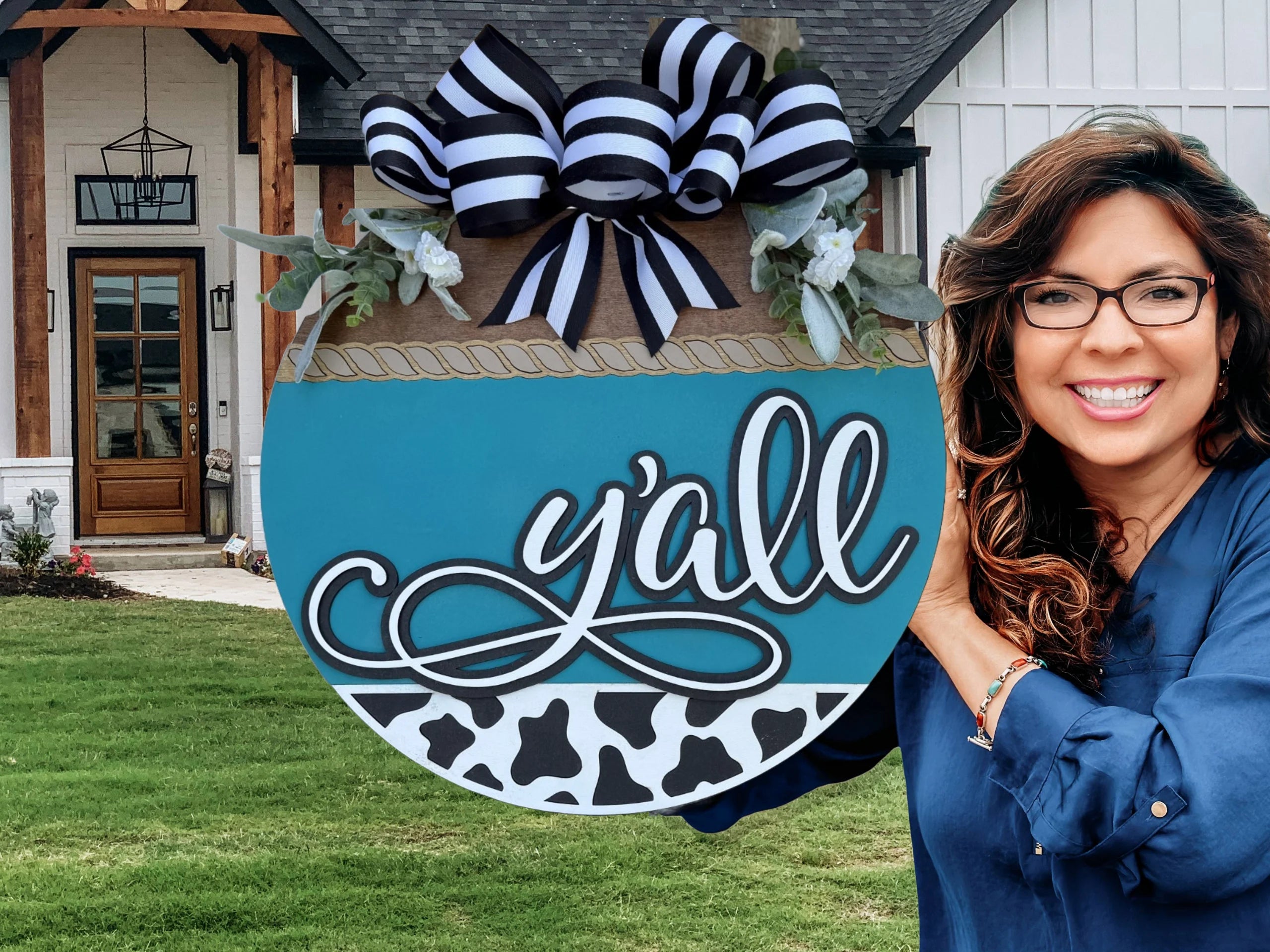 A smiling woman with wavy brown hair and glasses holds a round decorative sign that says yall in front of a modern farmhouse-style home with a wooden front door and manicured lawn.