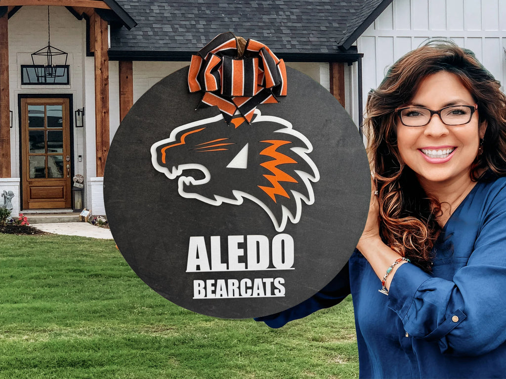 A smiling woman with long brown hair and glasses holds an Aledo Bearcat Mascot Door Hanger Door Sign, featuring the orange and black bearcat logo, while standing in front of a modern house with a green lawn.