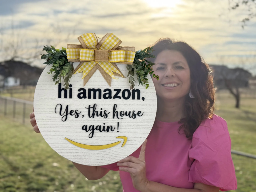 A woman in a pink shirt holds the Hi Amazon Yes This House Again Door Sign, decorated with green foliage and a yellow gingham bow, featuring a bright yellow arrow. In the background, theres a grassy field, trees, and a soft cloudy sky.