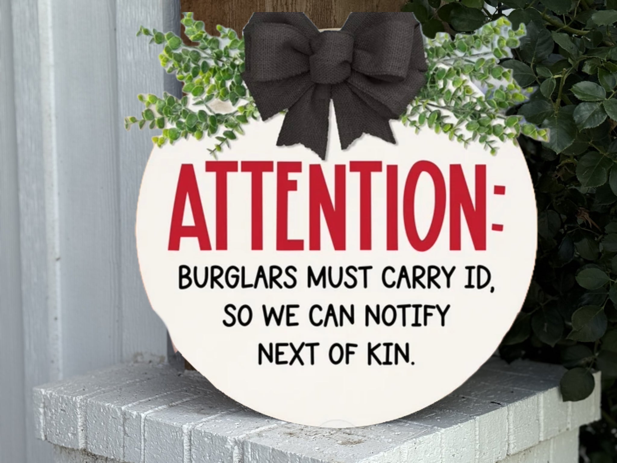 A round sign with a black bow and greenery reads: ATTENTION: BURGLARS MUST CARRY ID, SO WE CAN NOTIFY NEXT OF KIN. The sign sits on a white brick ledge beside a plant.