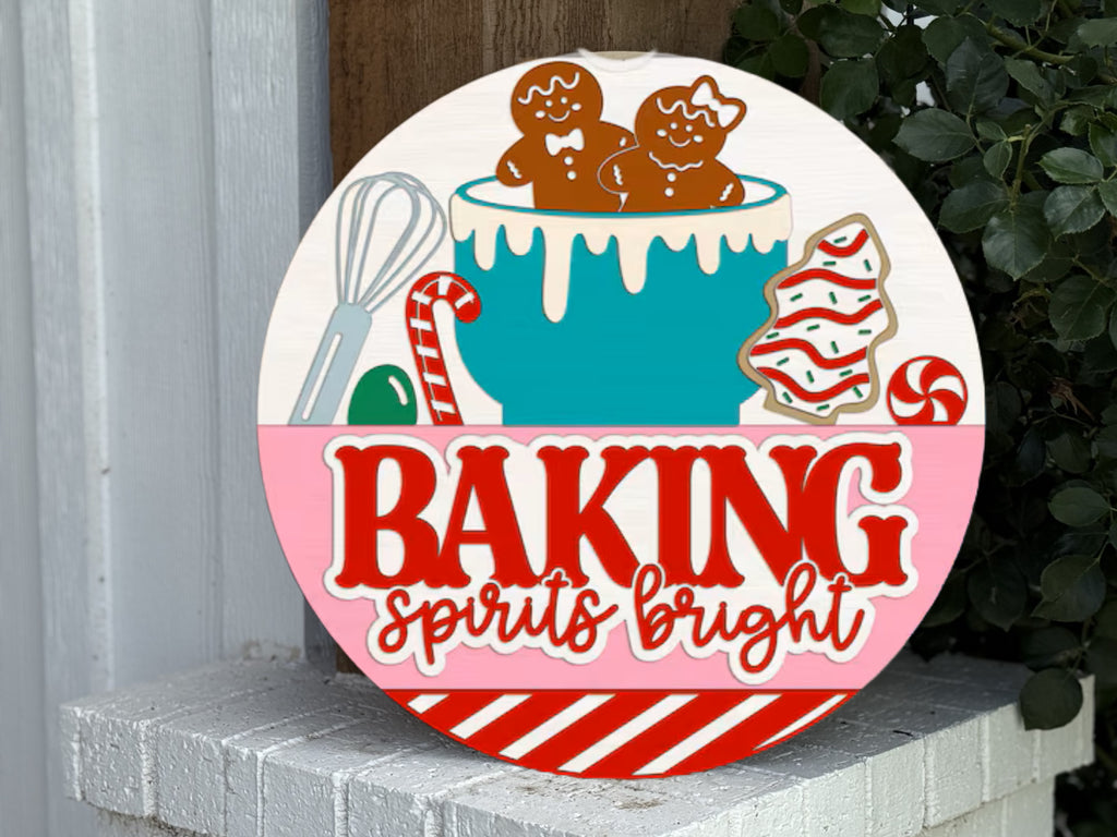 A festive sign with gingerbread cookies, icing, and baking tools in a bowl, decorated cookies, and the words Baking Spirits Bright in bold red and white letters. The sign rests on a white brick ledge near green leaves.