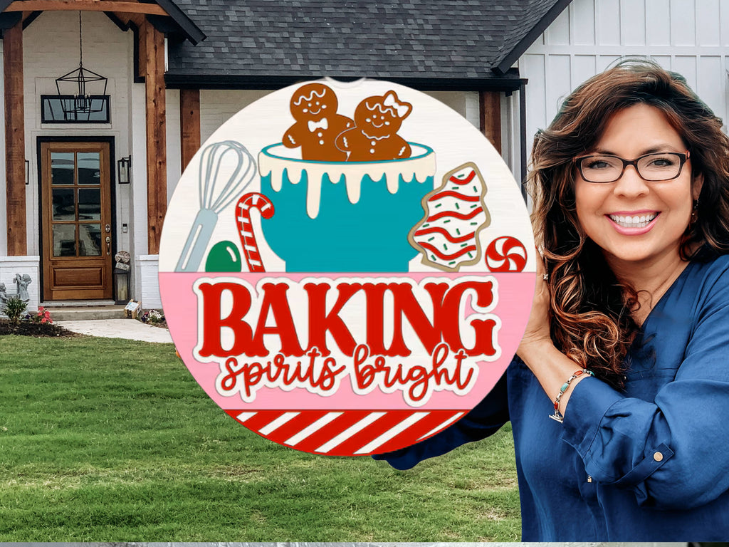 A smiling woman holds a festive sign with gingerbread cookies, candy canes, a whisk, and the words “BAKING spirits bright” in front of a modern house with a lawn and wooden accents.