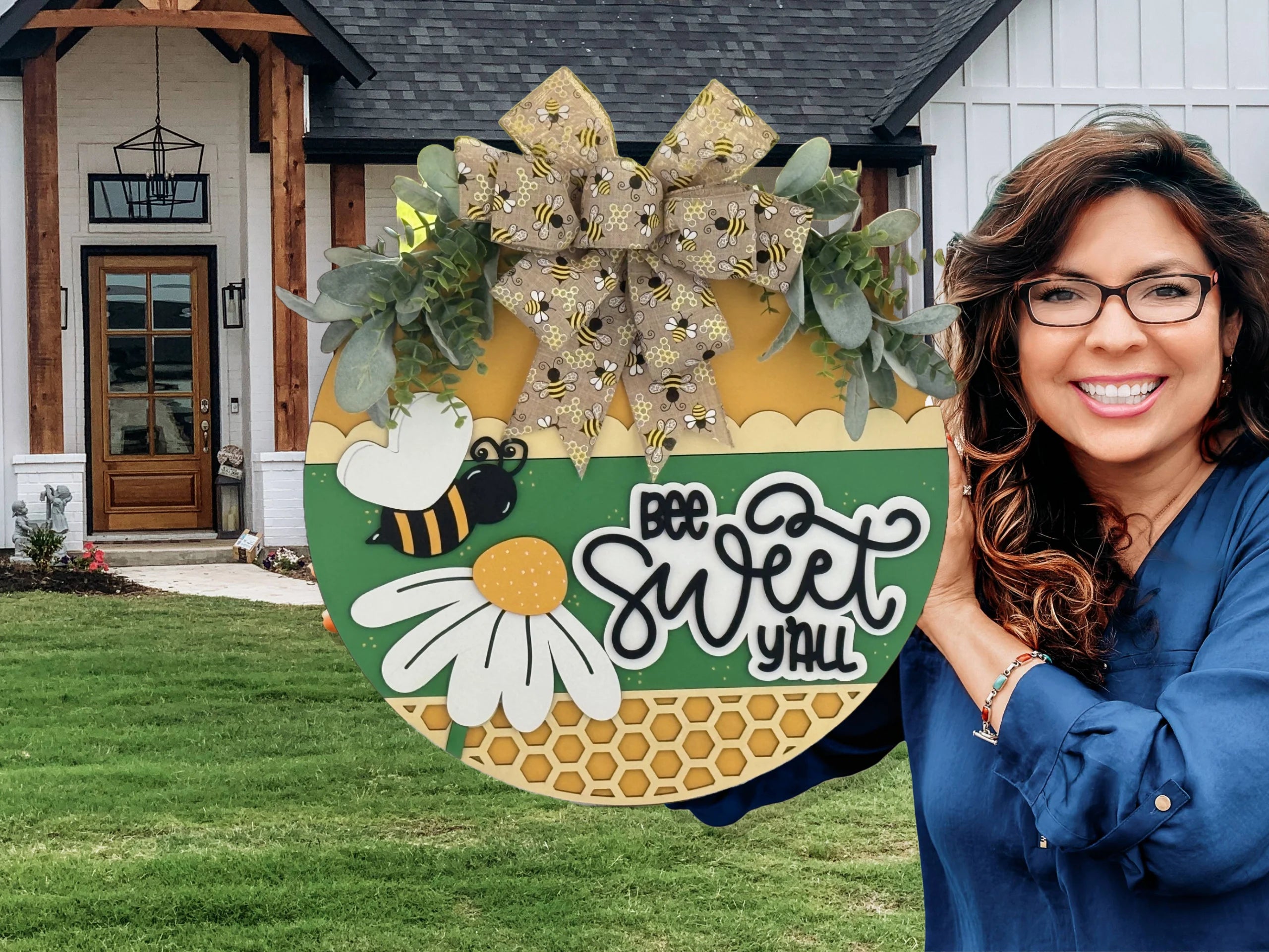 Woman in a blue shirt smiles while holding a circular, bee-themed decoration. The sign reads Bee Sweet Yall with a bee, daisy, and honeycomb design, and has a large bow and greenery on top. In the background, theres a modern house with wooden accents and a manicured lawn.