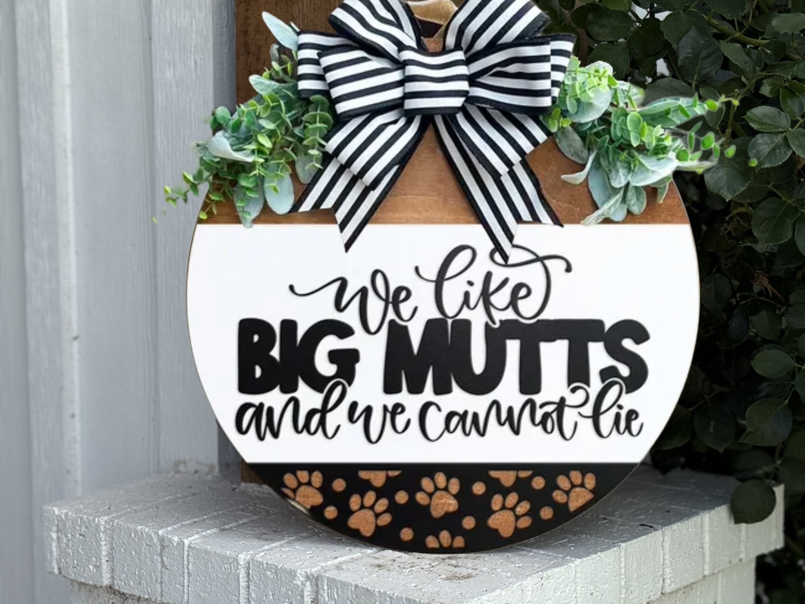 A round decorative sign with a black and white striped bow, greenery, and the text “We like BIG MUTTS and we cannot lie” above a strip with gold paw prints, displayed on a white brick surface.